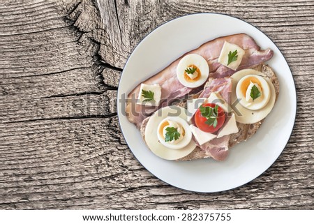 Toast Sandwich in White Porcelain Plate, with Pork Bacon Rasher, Ham, Edam and White Cow Cheese, hard boiled Egg slices, Cherry Tomato, Mayonnaise and Parsley leaves, on Old, Knotted, Wood surface.