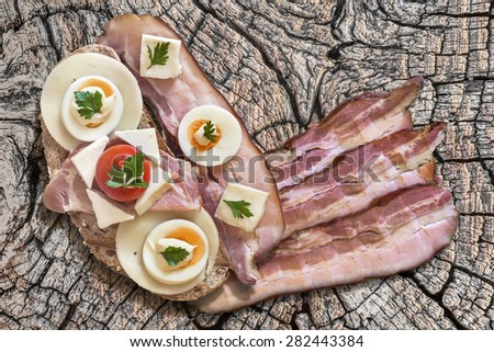 Toast Sandwich with Pork Bacon Rasher, and three extra Bacon Rashers, Ham, Edam and White Cow Cheese, hard boiled Egg slices, Cherry Tomato and Mayonnaise, on Old, Weathered, Cracked Wood surface.
