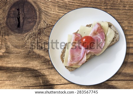 Plate with Sandwich made of slice of Integral Bread, with Pork Ham and Edam Cheese slices, with Cherry Tomato, with extra Belly Bacon rasher alongside, on old Wooden Table surface.