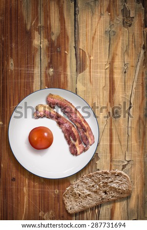 Photograph of Fried Bacon Rashers with Tomato on Porcelain Plate with Slice of Brown Integral Bread on old, peeled off, scratched Wooden Table.
