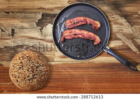 Fried Belly Bacon Rashers in Teflon Frying Pan with Bread Loaf alongside on old, cracked, scratched, peeled off, obsolete Wooden Table surface.