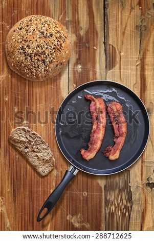 Fried Belly Bacon Rashers in Teflon Frying Pan with Bread slice and Loaf alongside on old, cracked, scratched, peeled off, obsolete Wooden Table surface.