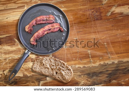 Fried Belly Bacon Rashers in Teflon Frying Pan with Integral Bread Slice alongside, on old, cracked, scratched, peeled off, obsolete Wooden Table surface.