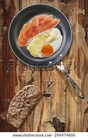 Fried Sunny Side Up Egg, with Edam Cheese and Cured Pork Ham Rashers, in heavy duty Teflon Frying Pan with Integral Bread Slice alongside, on Old, Cracked, Scratched, Peeled-off Wooden Table.