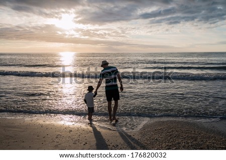 Father and son holding hands in front of the sunset at the beach wearing shorts and a hat
