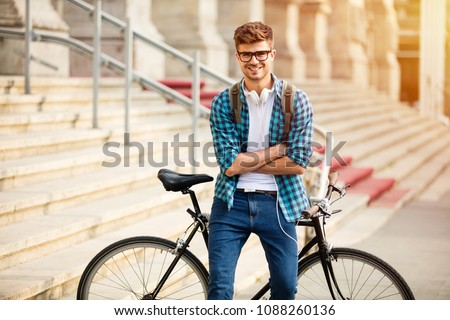 young student with glasses and bike standing on his bike on the street in front of university, looking at camera with confidence and big smile, with folded arms
