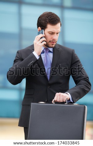 businessman talking on the phone in front of an office building with a briefcase in hand and looking at watch