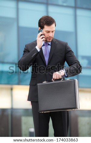 businessman talking on the phone in front of an office building with a briefcase in hand and looking at watch