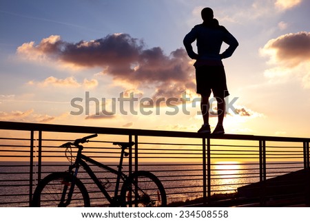 silhouette of young and active sportsman and his mountain bike standing on the railing near the ocean and looking far away at the sunset