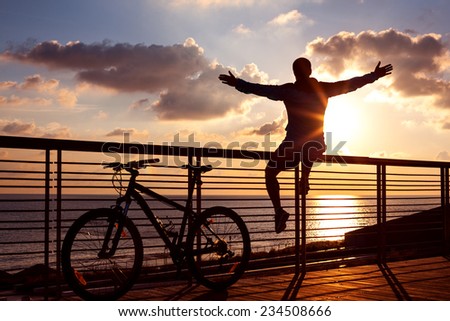 silhouette of young and active sportsman and his mountain bike sitting on the railing with outstretched arms near the ocean and looking far away at the sunset