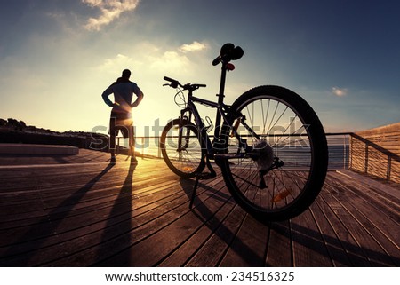silhouette of young and active sportsman and his mountain bike standing near the ocean and looking far away at the sunset