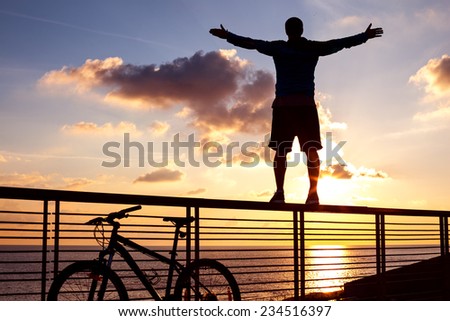 silhouette of young and active sportsman and his mountain bike standing on the railing with outstretched arms near the ocean and looking far away at the sunset