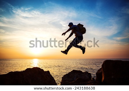 young brave man with backpack jumping over the rocks near the sea in sunset