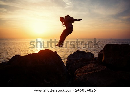brave man with backpack jumping over rocks near ocean in beautiful sunset