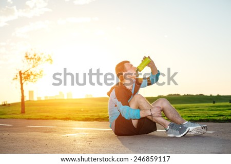sweaty tired runner sitting in the park on the road and drinking water from sport bottle