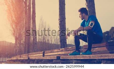 Tired athlete resting on the bench in park with bottle of water, armband with mobile phone and listening music (intentional sun glare and vintage color)