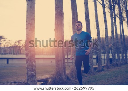 Handsome athlete running in the park along trees at sunset (little motion blur, intentional sun glare and vintage color)