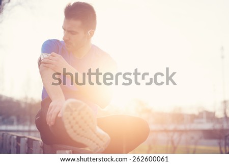 Athlete sitting in the park at sunset touching armband with mobile phone (intentional sun glare and vintage color)