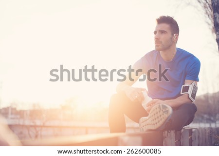 Athlete resting on the bench in park with bottle of water, armband with mobile phone and listening music (intentional sun glare and vintage color)