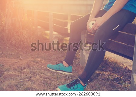 Tired athlete with bottle of water resting near bench in park (intentional sun glare and vintage color)