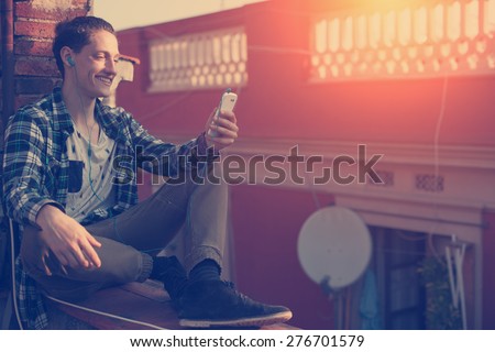 Young and smiling man sitting on the roof with mobile phone and listening music (intentional sun glare and vintage color, focus on mobile phone)