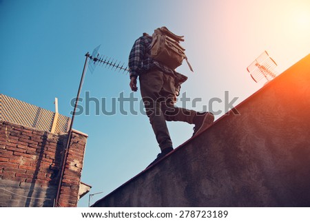Brave man with backpack walking over high wall on the edge, on the rooftop (intentional sun glare)