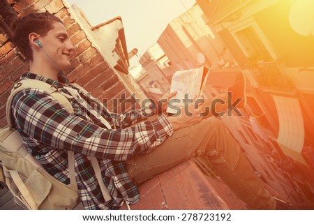 Young and brave man with backpack sitting on the edge of high roof with map in hands (intentional sun glare)