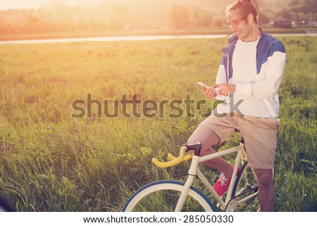 young man with bicycle in green field looking at smartphone at sunset (intentional sun glare and vintage colors)