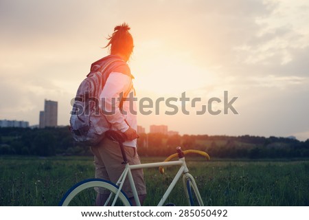 young man with bicycle in the green field looking far away at sunset (intentional sun glare and dark colors)