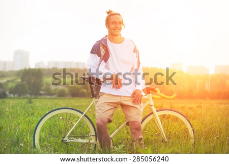Man in blank t-shirt with bicycle standing in green field at sunset (intentional sun glare and bright color)