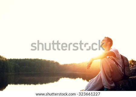 Young and handsome tourist sitting near the lake at sunset (intentional sun glare and lens flares)