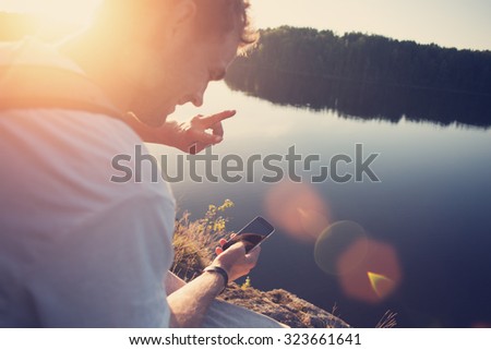 Young happy traveler making selfie with mobile phone near the lake outdoors (intentional sun glare and lens flares)