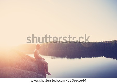 Young man sitting on the edge of the cliff near the lake after swimming and looking far away (intentional sun glare, lens flares and vintage color)