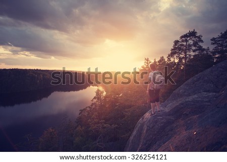 Lonely traveler man with backpack looking at sunset near the river (intentional sun glare and lens flare)