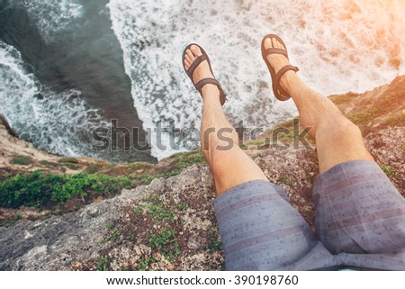 Young brave man sitting on a high cliff above ocean (intentional sun glare, POV view)