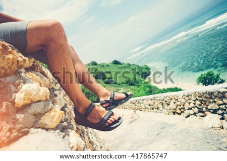 Man\'s legs sitting on the rocks above the road near ocean. Intentional sun glare