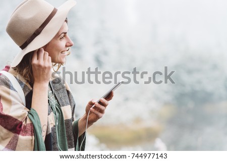 Smiling handsome woman listening music with her mobile phone in first snow. Traveling among stunning winter landscape. Vacations in mountain wilderness. Wanderlust and boho style