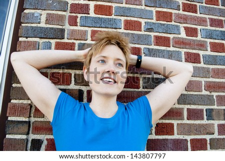 A young woman smiling again a brick wall with arms wrapped behind her head
