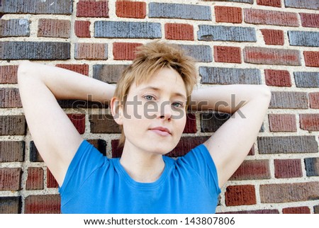 An androgynous young woman looking at the camera with arms behind her head, leaning against a brick wall