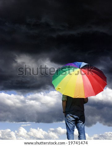 A person standing alone, watching a storm approach, with a rainbow coloured umbrella.
