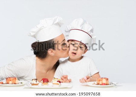 Cook chef mother kissing her son on cheek w,th bunch of sweet dessert dishes