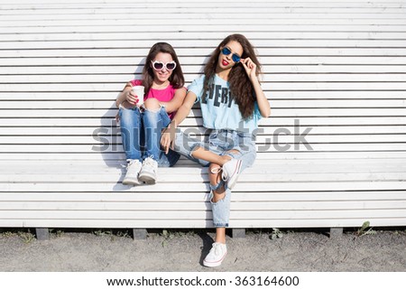 Two young girls. Portrait of two pretty hipster sitting on a bench, resting in the sun. Girls smile, have fun. Outdoor lifestyle portrait. Girl with glasses drinking coffee.