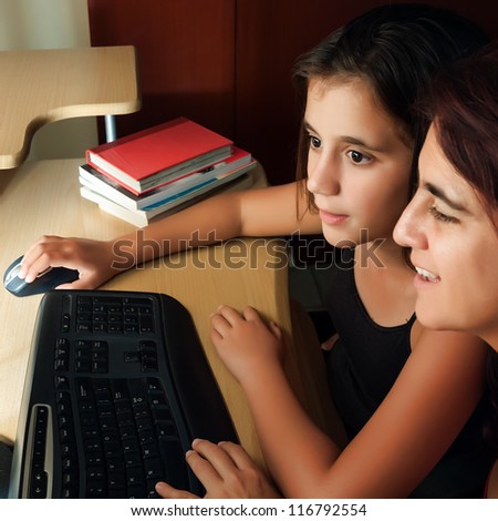 Hispanic mother and daughter browsing the web at home with the light from the computer monitor illuminating their faces (with books on the computer desk)