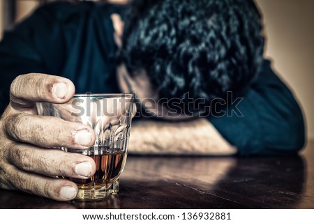 Depressed drunk man holding a drink and sleeping with his head on the table (Focused on the drink, his face is out of focus)