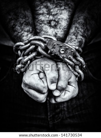 Dramatic black and white image of the hands of a shirtless  man chained with an iron chain and a padlock (emerging from a black background)