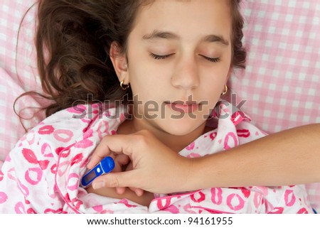 Cute hispanic girl sick with fever laying in her bed while a woman\'s hand measures her temperature with a thermometer