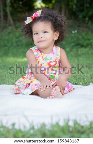 Funny small latin girl with a colorful dress sitting on a green grass park