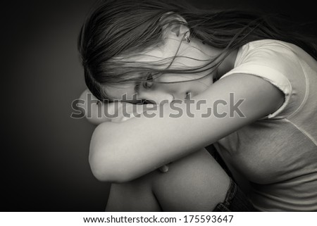 Black and white image of a sad and lonely girl with her head resting on her legs and looking at the camera