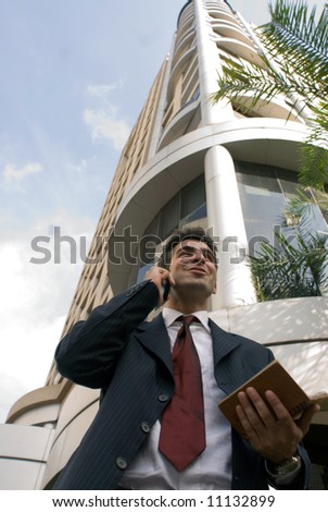A business man standing in front of a building, holding a day planner, talking on his cellphone.