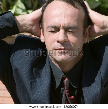 A shot of a business man taking in the Sun outdoors.  His hand are resting on the back of his head and his eyes are closed.